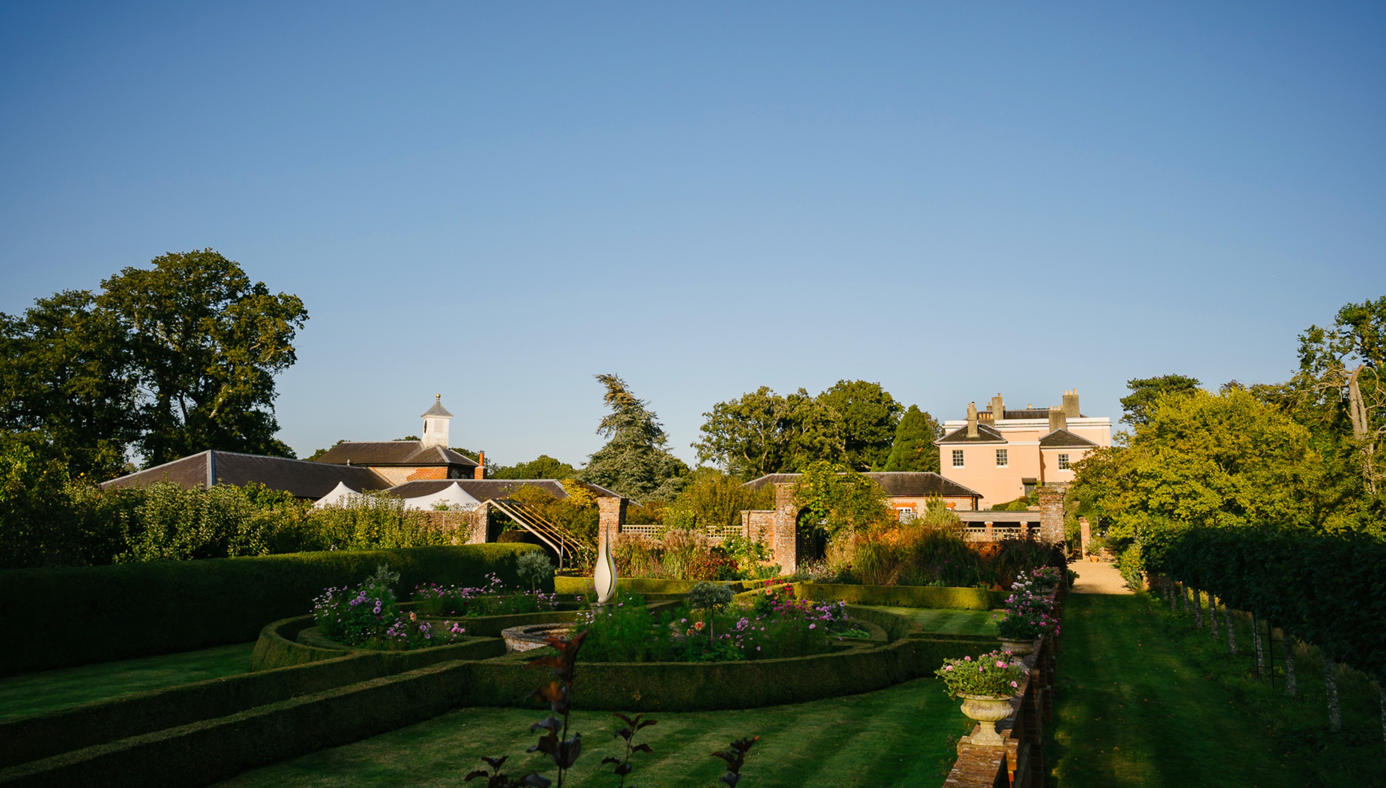 Wide view of Bignor Park’s formal gardens at golden hour, with sculpted hedging, flowering borders and the house beyond in the West Sussex countryside