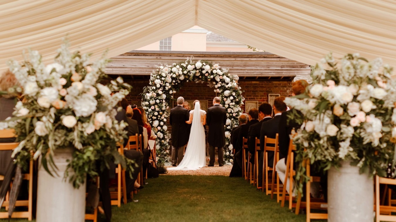 Wedding ceremony inside marquee at Bignor Park in West Sussex with floral arch and aisle seating