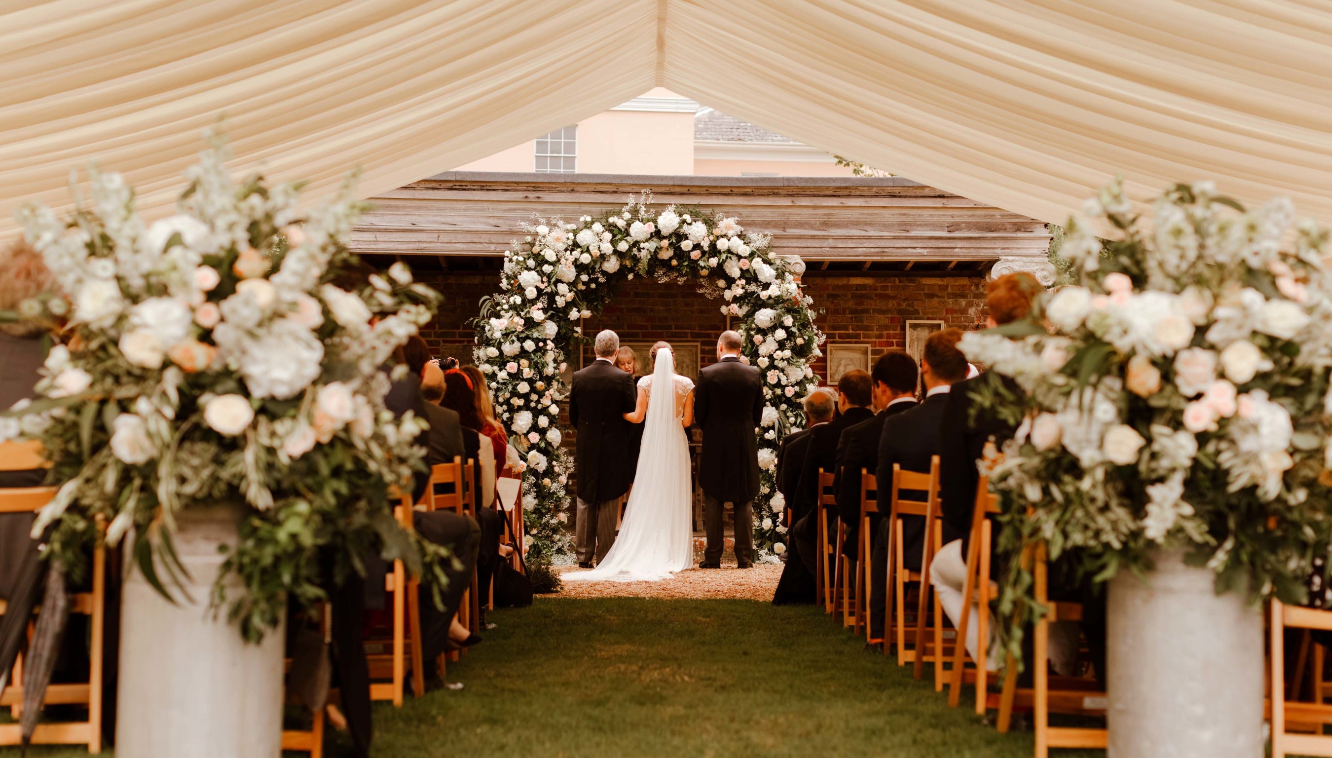 Wedding ceremony inside marquee at Bignor Park in West Sussex with floral arch and aisle seating
