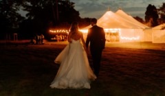 Bride and groom walking towards a lit marquee on the Croquet Lawn Bignor Park in the evening with festoon lights glowing