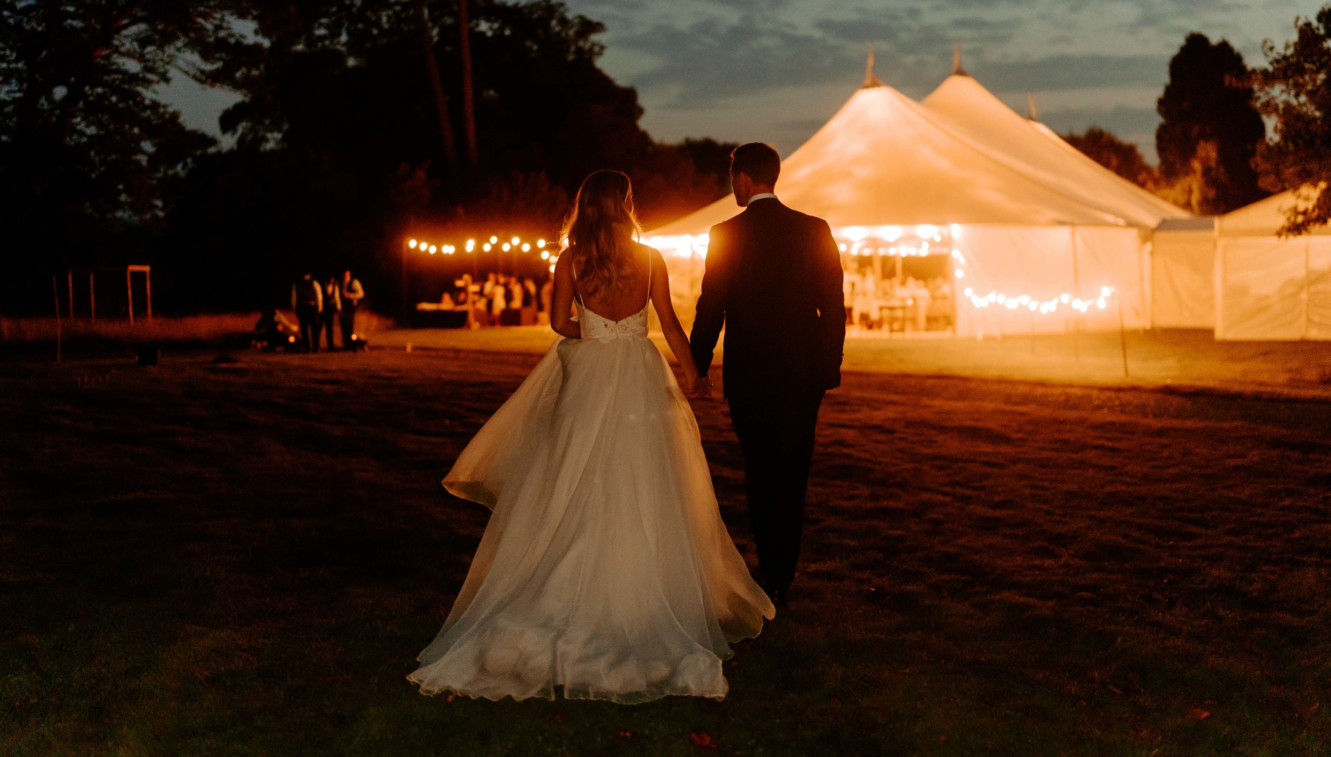 Bride and groom walking towards a lit marquee on the Croquet Lawn Bignor Park in the evening with festoon lights glowing
