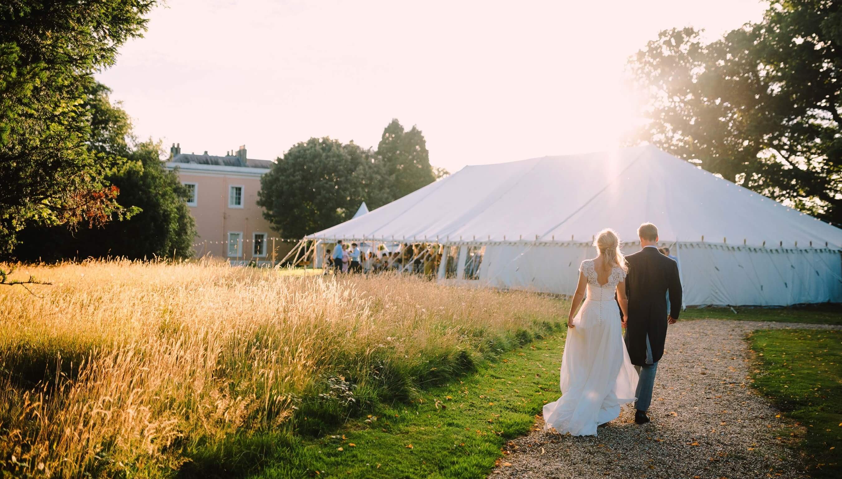 Bride and groom walking through Bignor Park grounds in West Sussex towards wedding marquee on the Croquet lawn at sunset