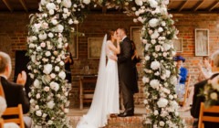 Bride and groom kissing during wedding ceremony at Bignor Park in West Sussex beneath a floral arch at the Greek Loggia