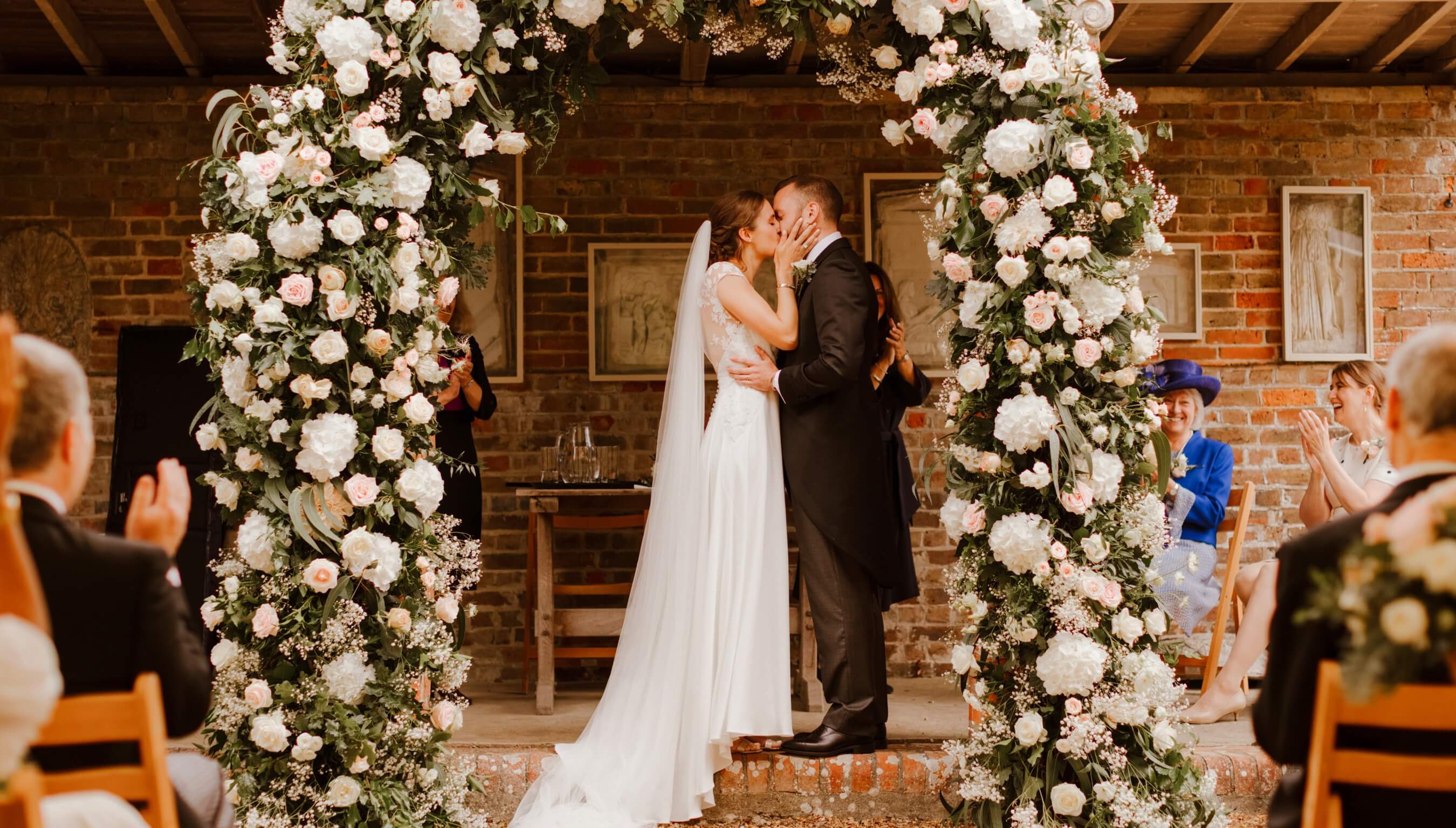 Bride and groom kissing during wedding ceremony at Bignor Park in West Sussex beneath a floral arch at the Greek Loggia