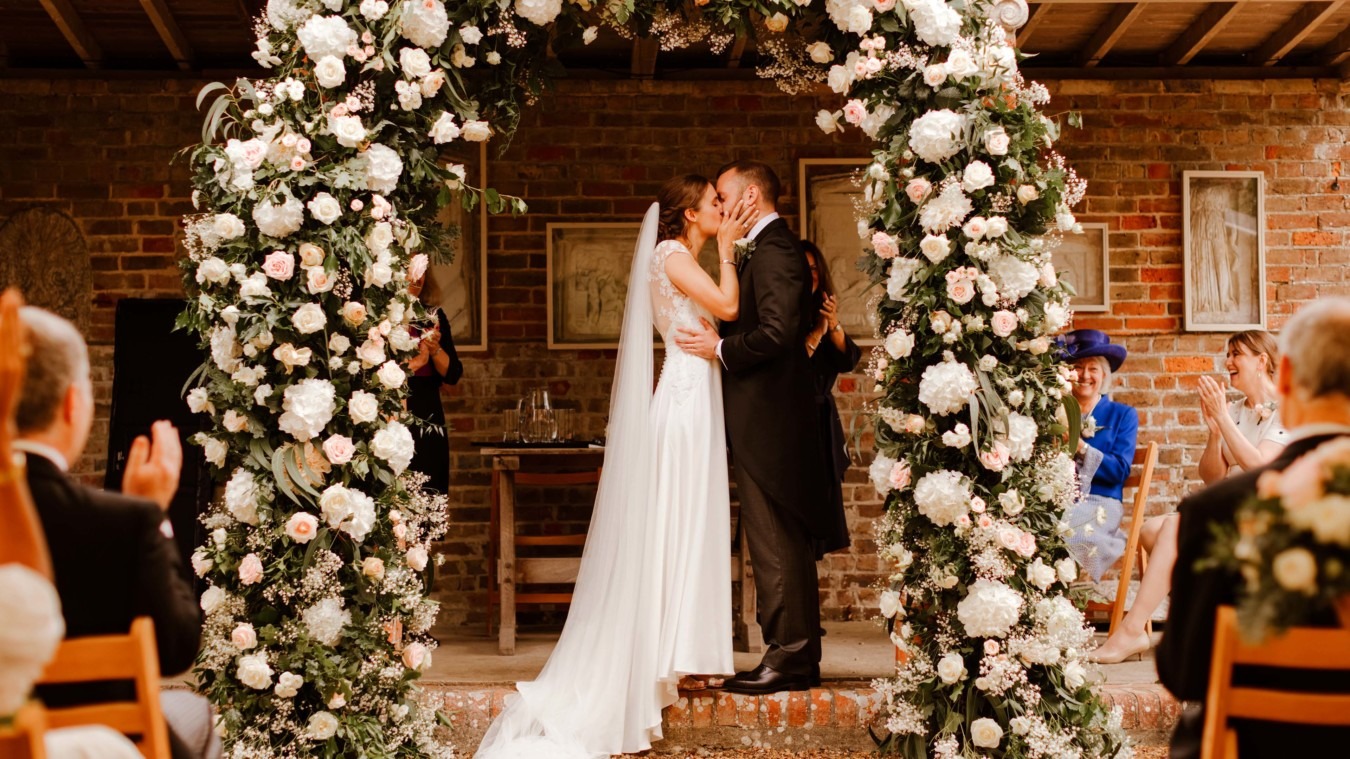 Bride and groom kissing at the Greek Loggia ceremony at Bignor Park in West Sussex with floral arch
