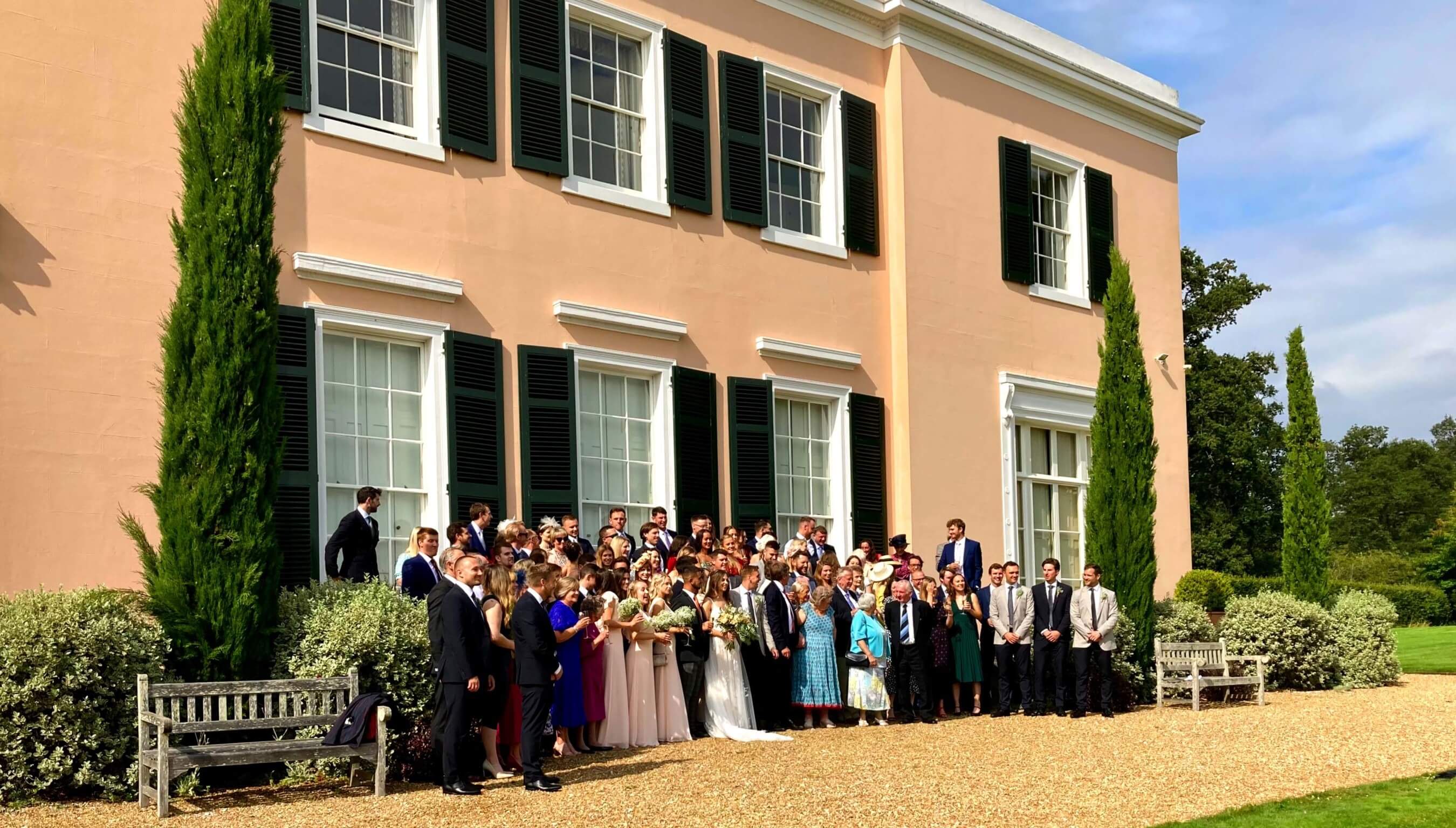 Large wedding group photo outside Bignor Park country house with guests gathered in front of peach facade and green shutters