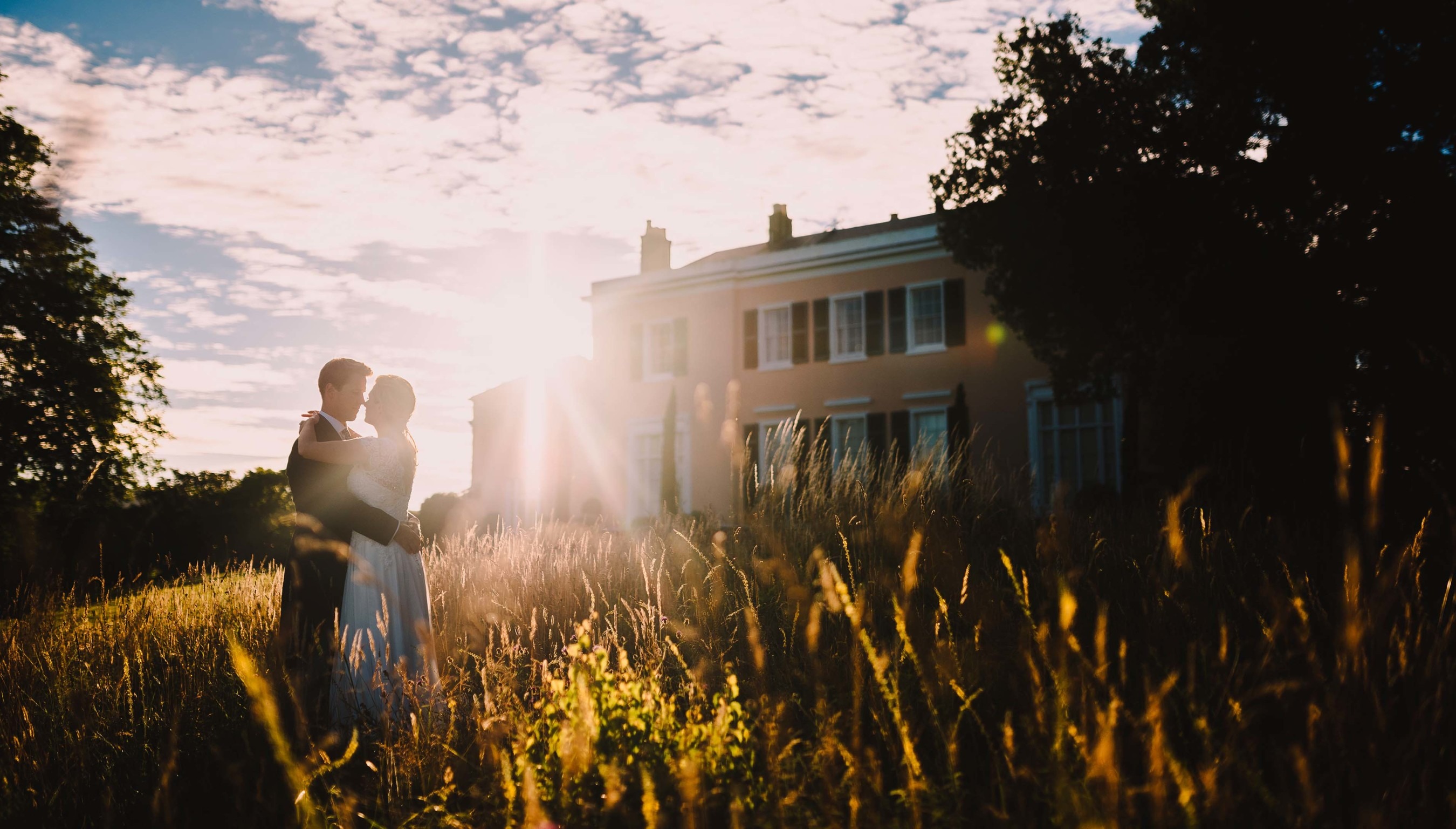 Bride and groom embracing at sunset in front of Bignor Park house in West Sussex, surrounded by wild meadow grass