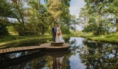 Bride and groom standing on wooden platform over reflective garden pond surrounded by trees at Bignor Park West Sussex