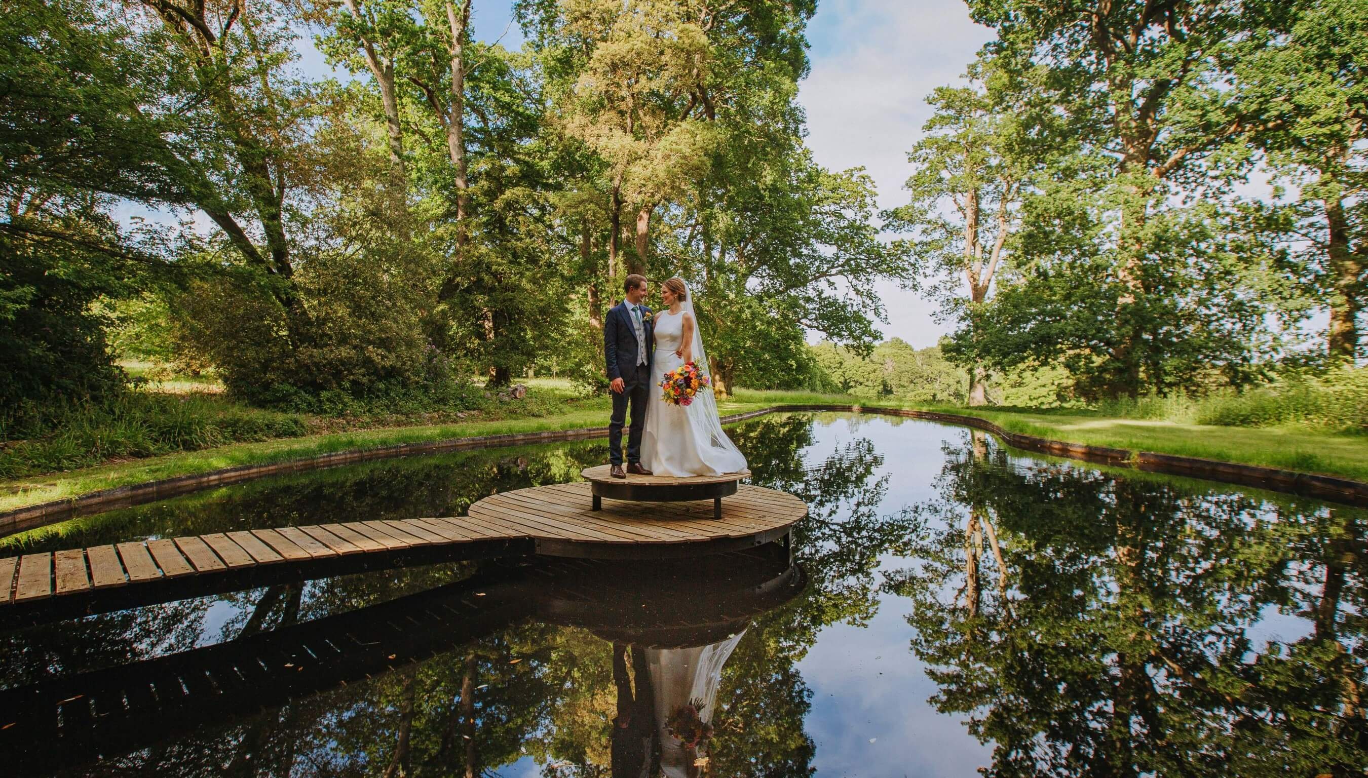 Bride and groom standing on wooden platform over reflective garden pond surrounded by trees at Bignor Park West Sussex