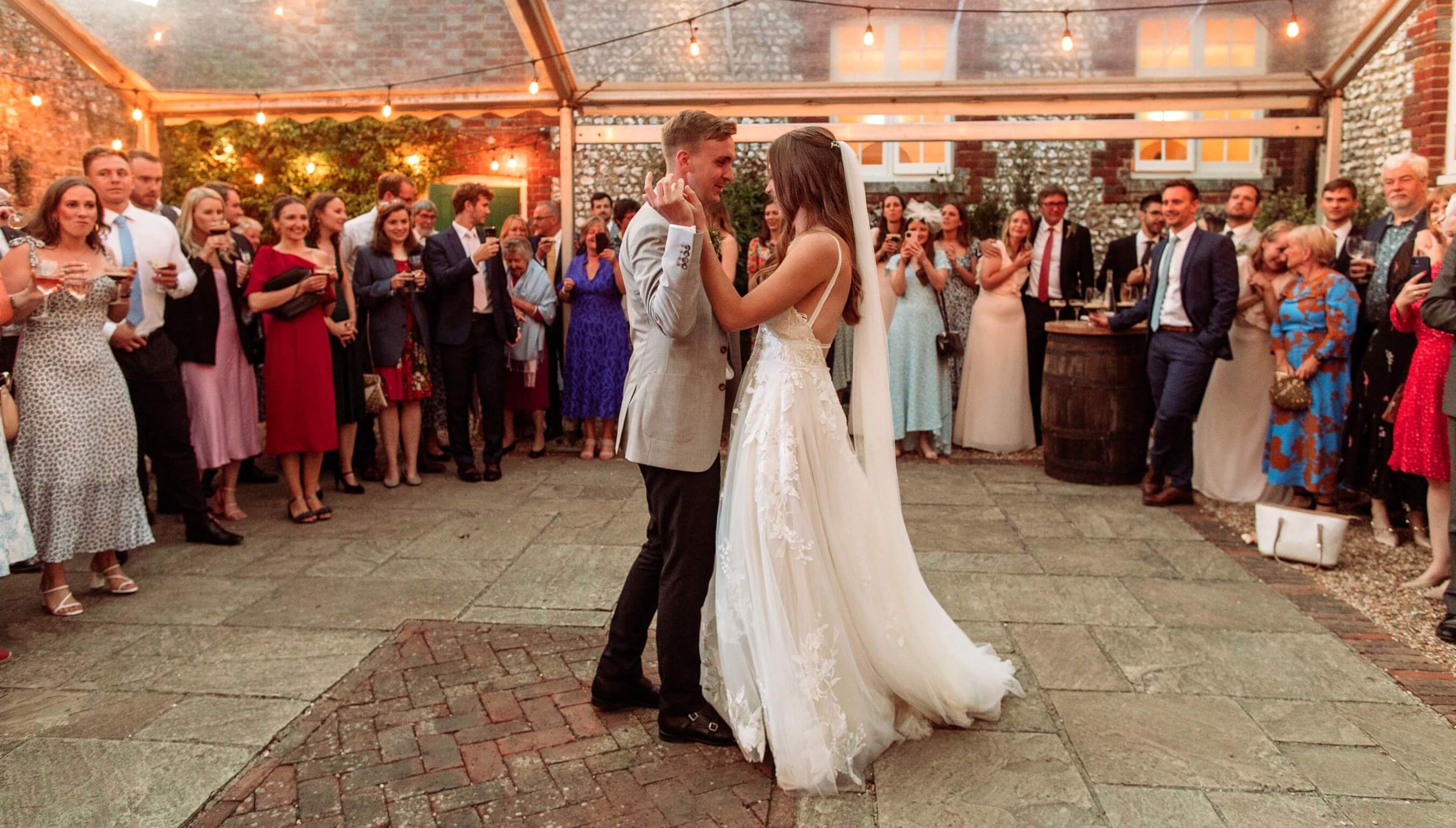 Bride and groom sharing their first dance in the Stables Quadrangle at Bignor Park with guests gathered around under festoon lighting