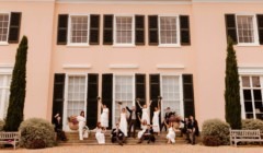 Wedding party celebrating on the steps of Bignor Park House with bride, groom, bridesmaids and groomsmen raising bouquets