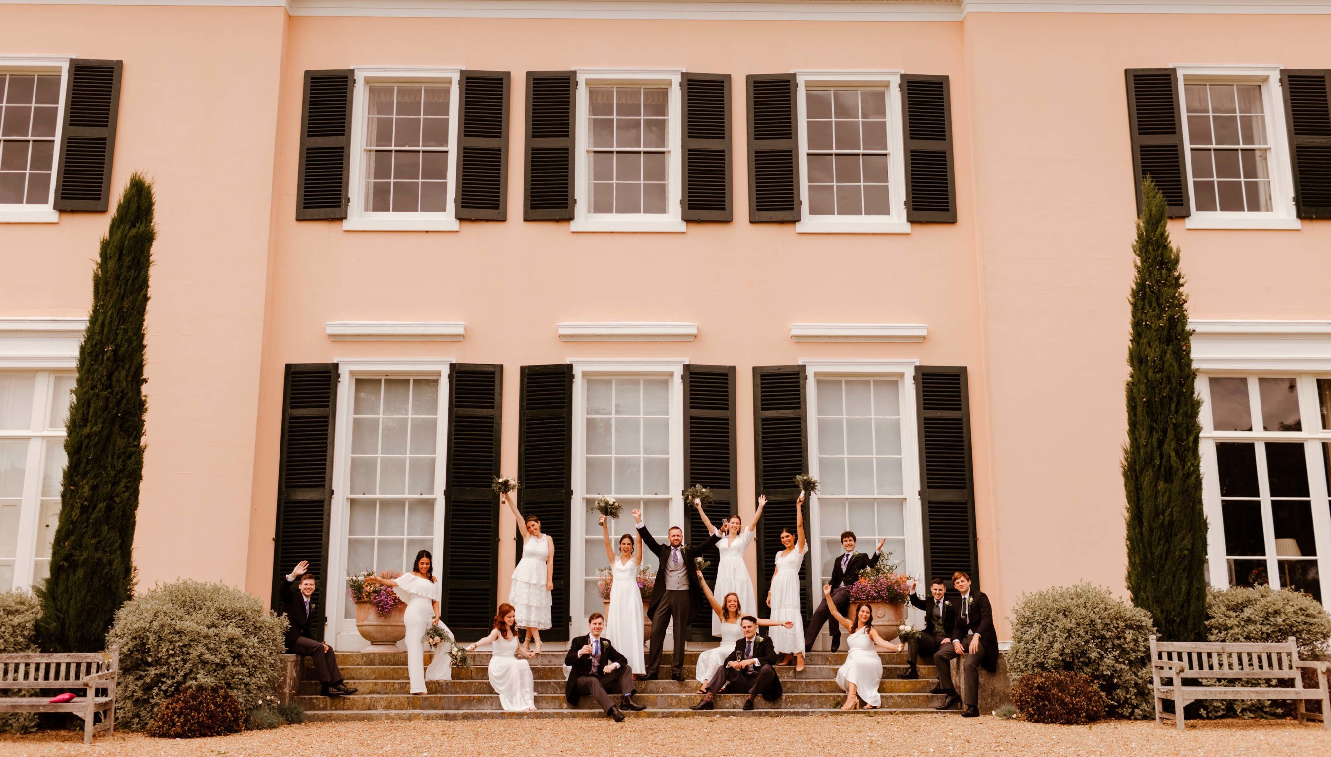 Wedding party celebrating on the steps of Bignor Park House with bride, groom, bridesmaids and groomsmen raising bouquets