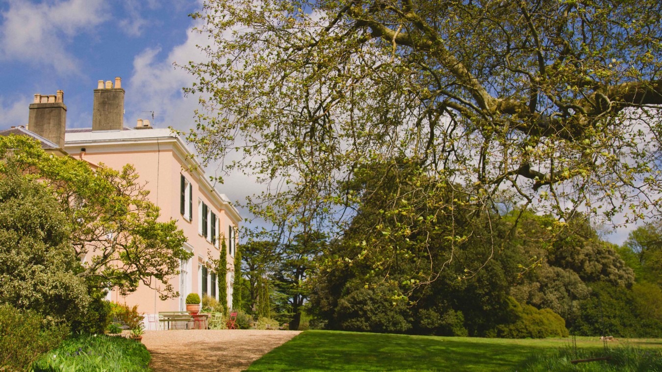 Elegant Georgian country house at Bignor Park surrounded by landscaped gardens, mature trees, and sweeping lawns under a bright spring sky