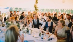 Wedding guests laughing and reacting during speeches at a marquee reception at Bignor Park, seated at decorated tables with drinks and floral centrepieces