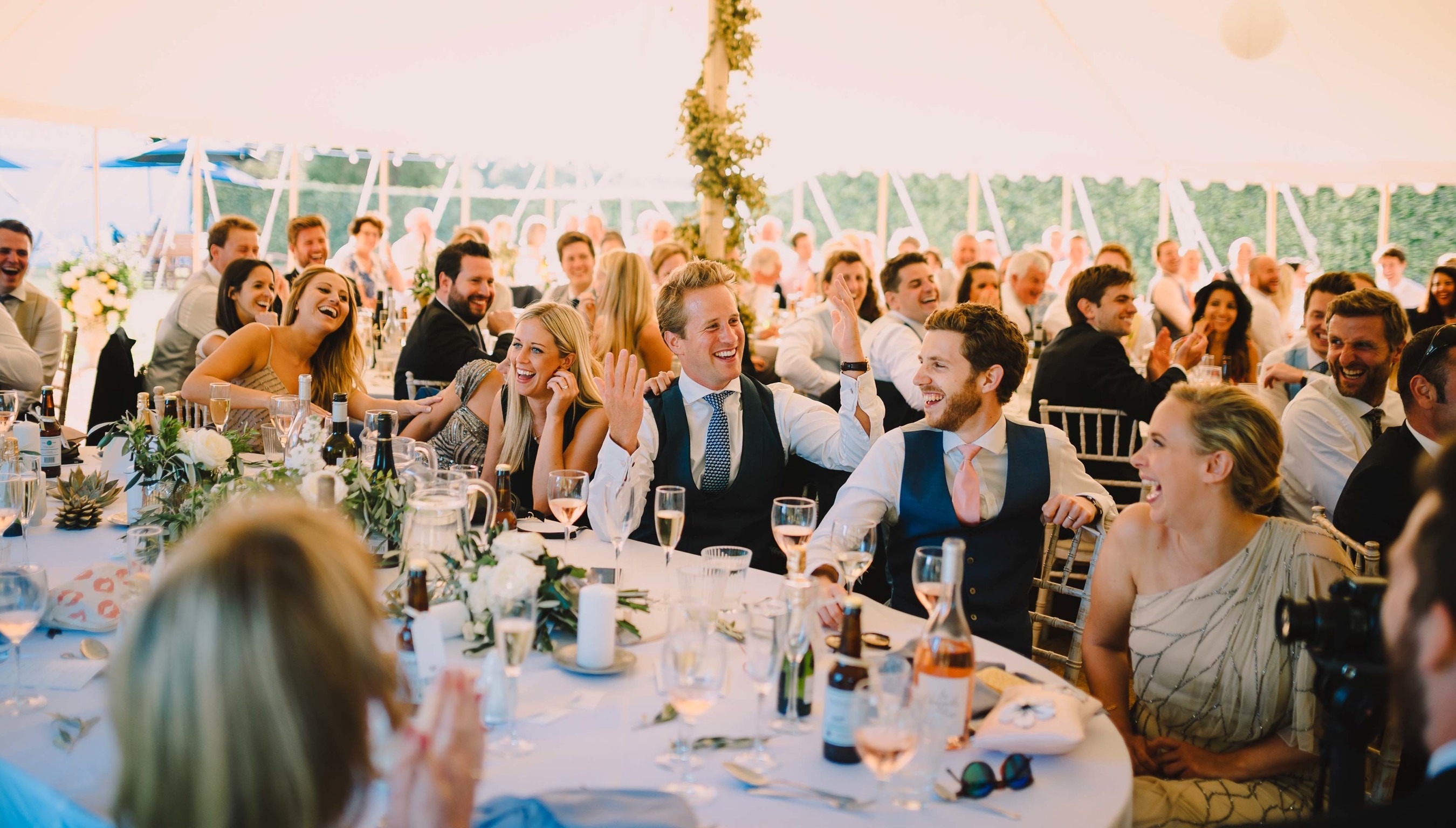 Wedding guests laughing and reacting during speeches at a marquee reception at Bignor Park, seated at decorated tables with drinks and floral centrepieces