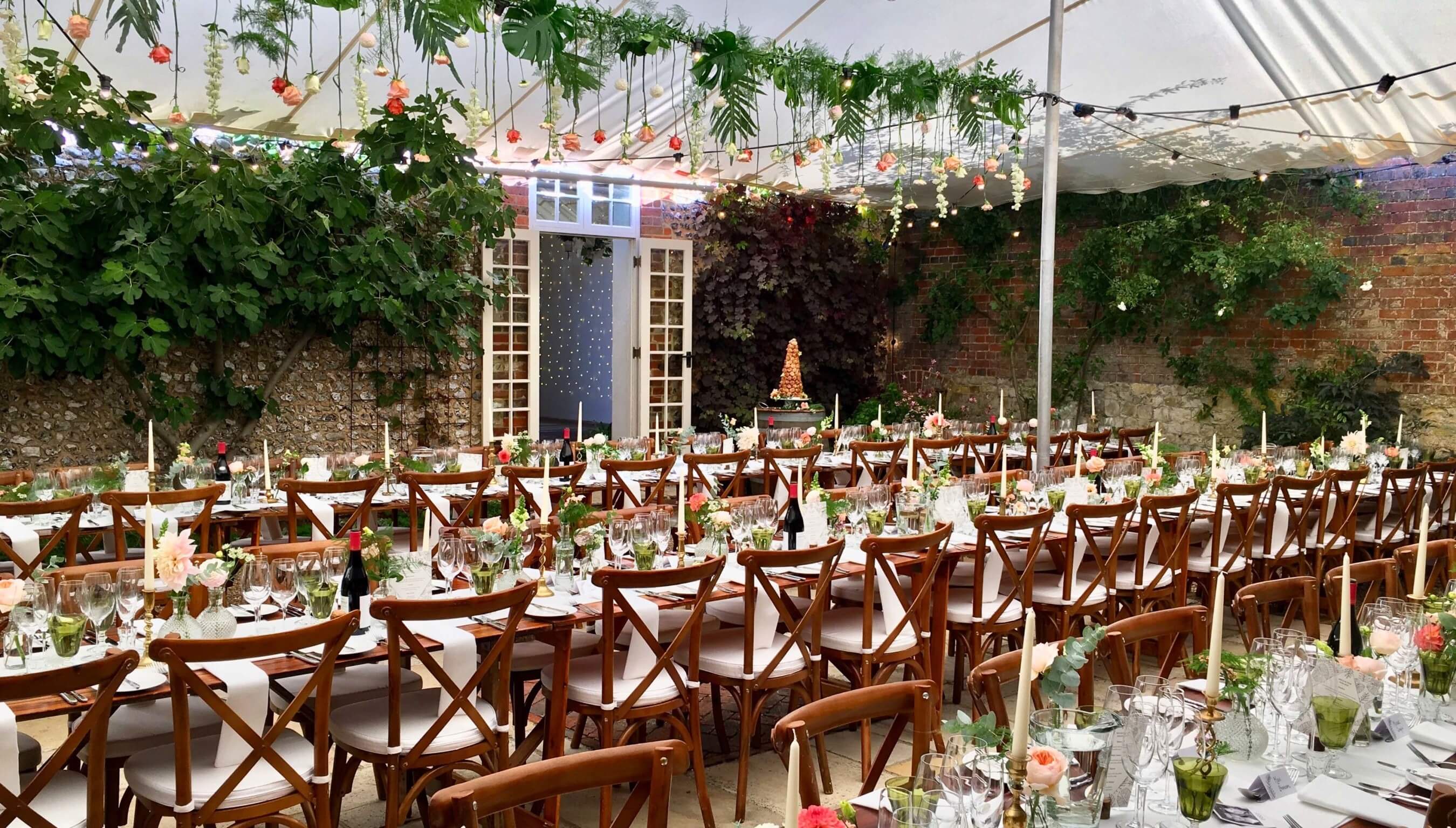 Wedding reception tables set in courtyard marquee at Bignor Park West Sussex with festoon lighting and floral décor