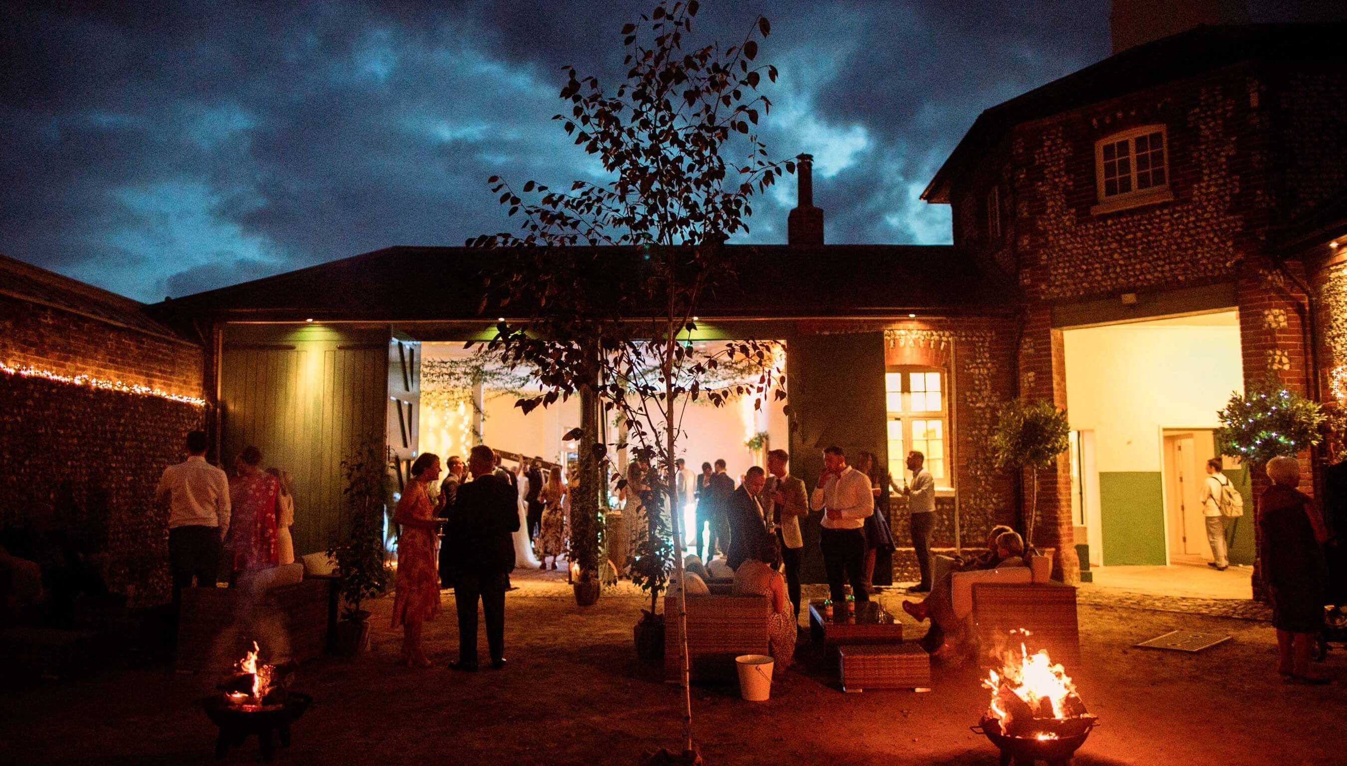Evening wedding reception at Bignor Park West Sussex with courtyard fire pits, festoon lighting and guests socialising outdoors