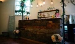 Rustic wedding bar setup at Bignor Park with reclaimed wood counter, hanging lights, greenery decor and drinks display inside The Stables reception space