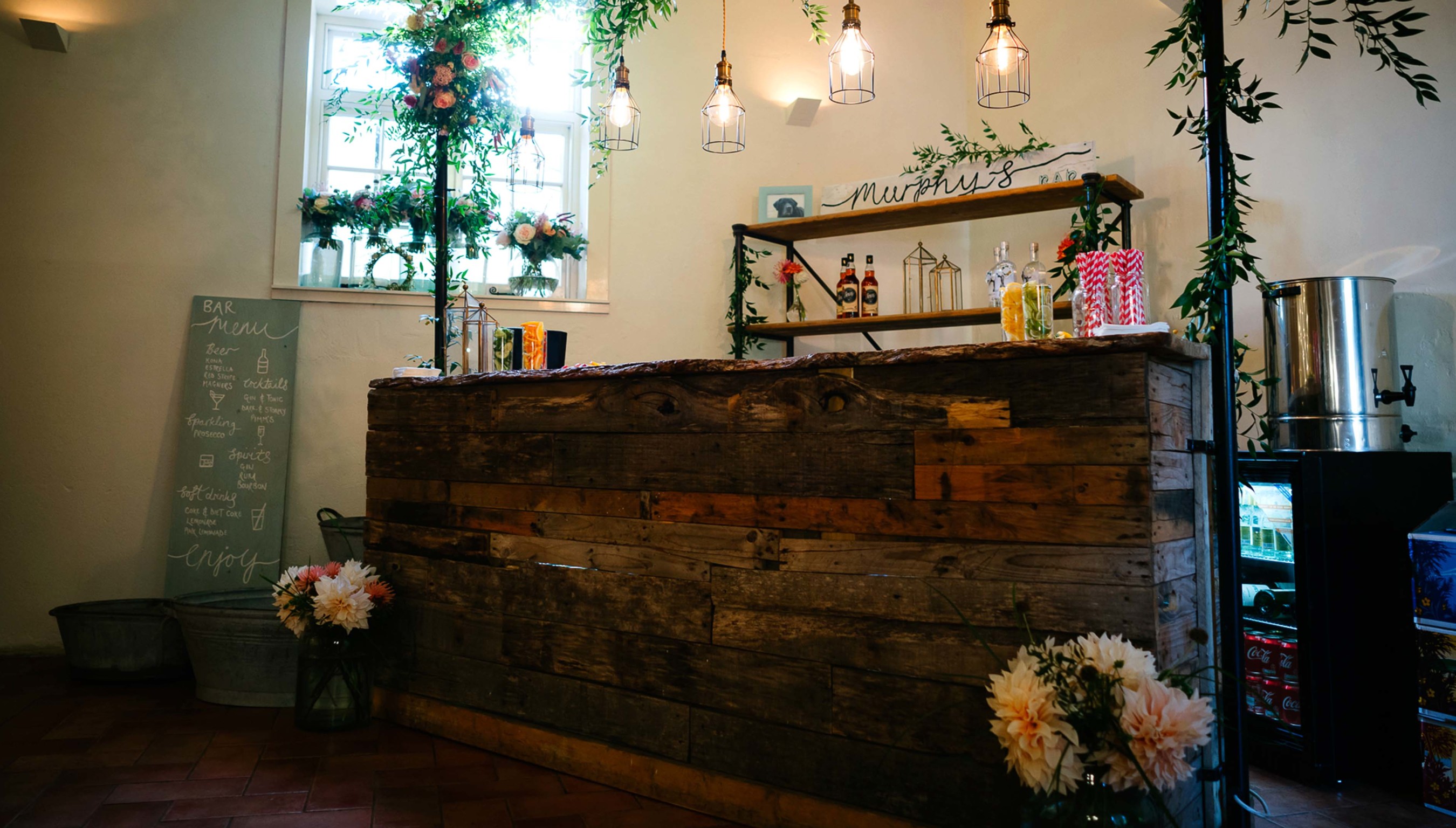 Rustic wedding bar setup at Bignor Park with reclaimed wood counter, hanging lights, greenery decor and drinks display inside The Stables reception space