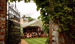 Outdoor wedding ceremony at Bignor Park’s Greek Loggia, viewed through an ornate iron gate with guests seated beneath a white canopy and the house beyond