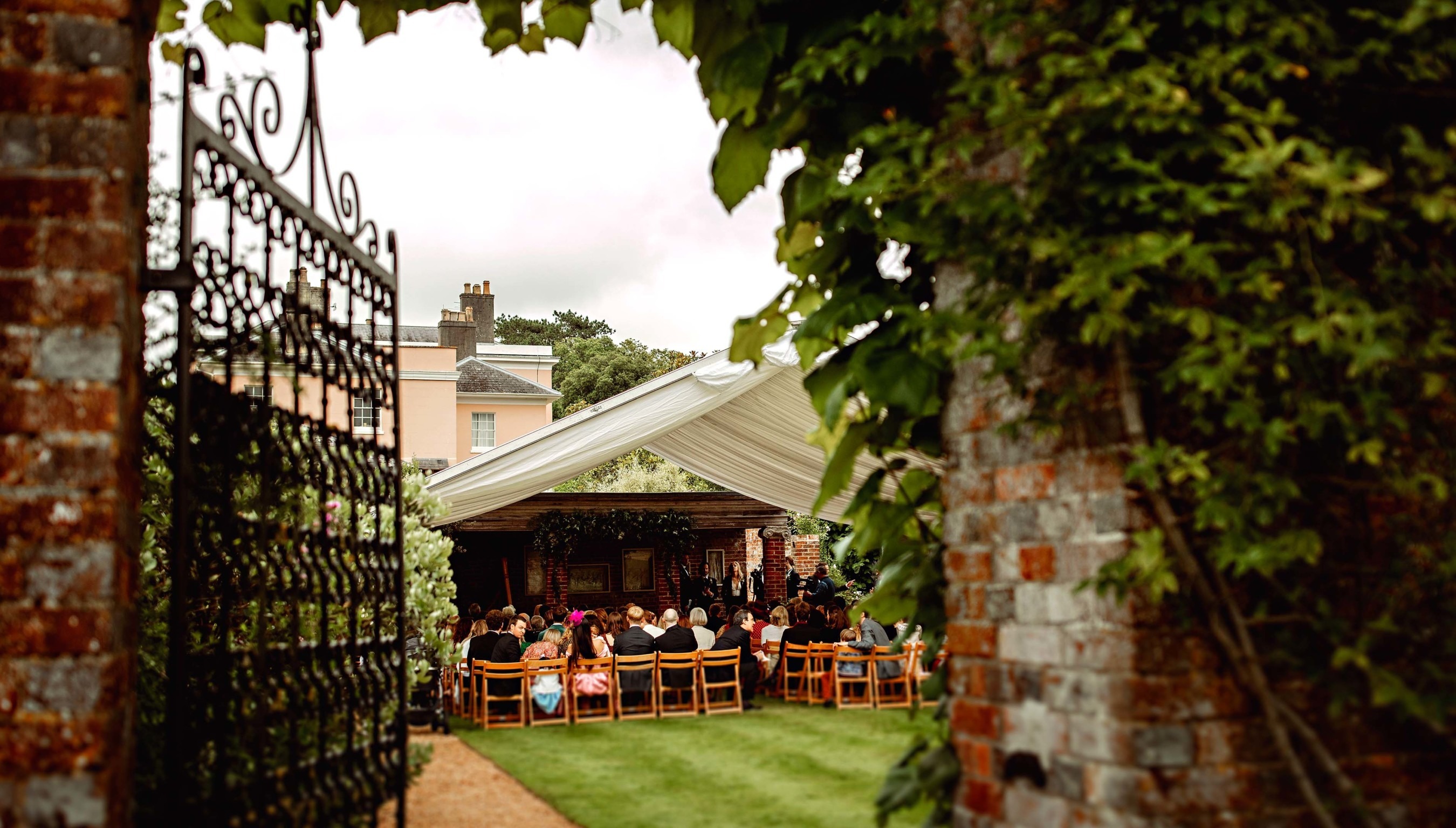 Outdoor wedding ceremony at Bignor Park’s Greek Loggia, viewed through an ornate iron gate with guests seated beneath a white canopy and the house beyond