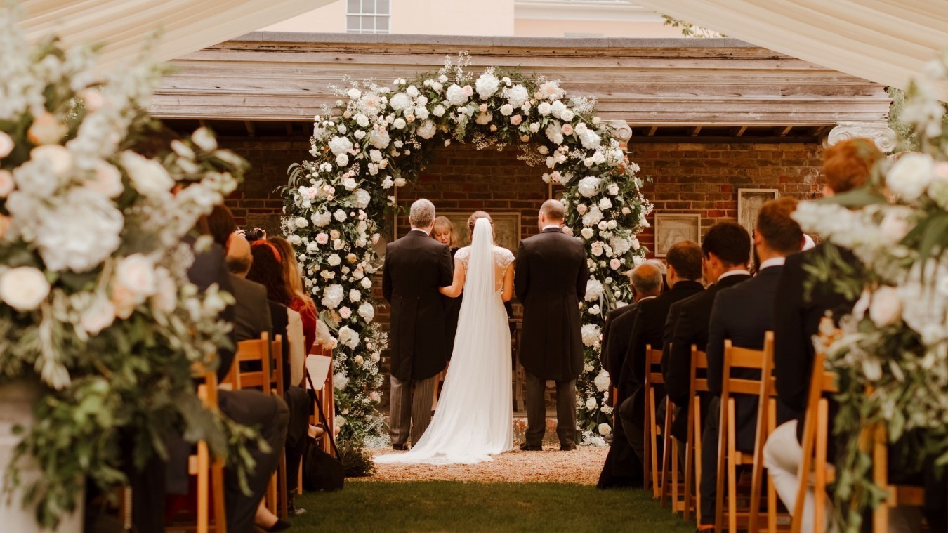 Wedding ceremony inside marquee at Bignor Park in West Sussex with floral arch and aisle seating