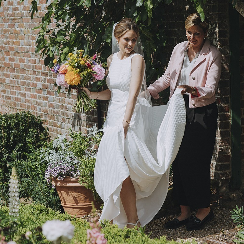 Bride in a simple white gown stepping through a garden doorway at Bignor Park, assisted by Louise Hartley, with colourful flowers and brick walls surrounding them