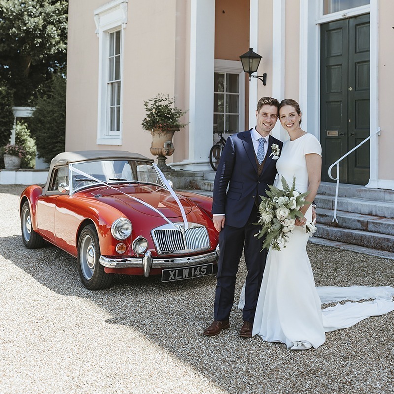 Wedding couple in front of Bignor Park country house in West Sussex with red vintage car